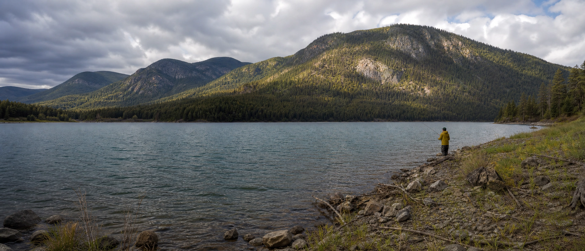 A lone lure angler works the rocky shore of a mountain lake beneath broken clouds